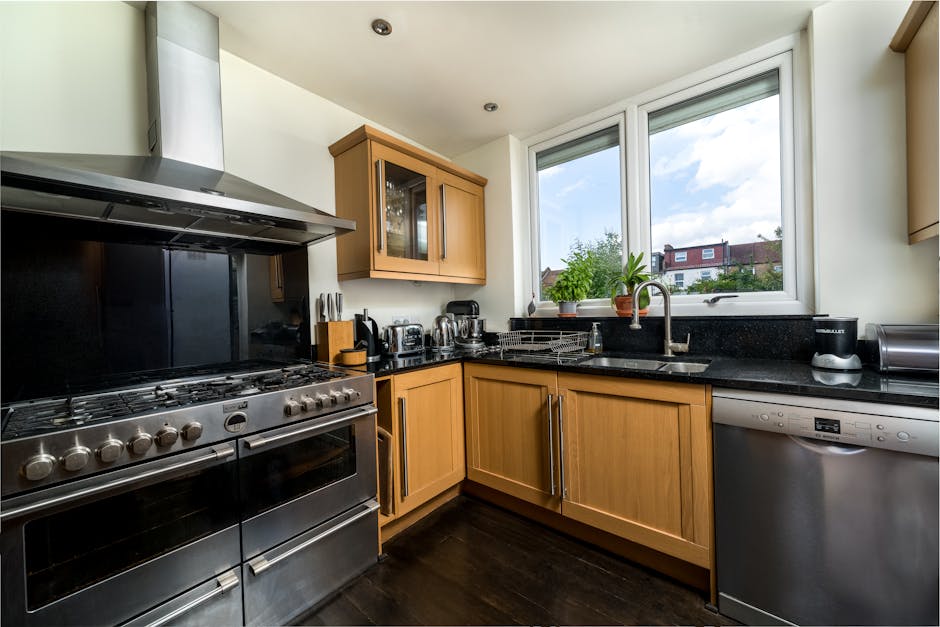 A modern kitchen with wooden cabinetry and black granite countertops illuminated by natural light from a large window overlooking residential buildings and greenery outside. The stainless steel oven and stove are clean, with the stovetop free of spills. On the countertop near the window, there is a stainless steel sink with a chrome faucet, beside a potted plant and various small appliances including a toaster, kettle, and coffee maker, all appearing well-maintained and dust-free. The kitchen surfaces, including the ceramic tiled backsplash behind the stove and the dark wood flooring, are spotless, reflecting a recent deep cleaning by Deep Cleaning Highbury to ensure hygiene and shine. The overall appearance emphasizes a tidy, hygienic environment suitable for thorough surface cleaning and sanitisation, aligned with our domestic cleaning services for Highbury Barn and N5 estates.