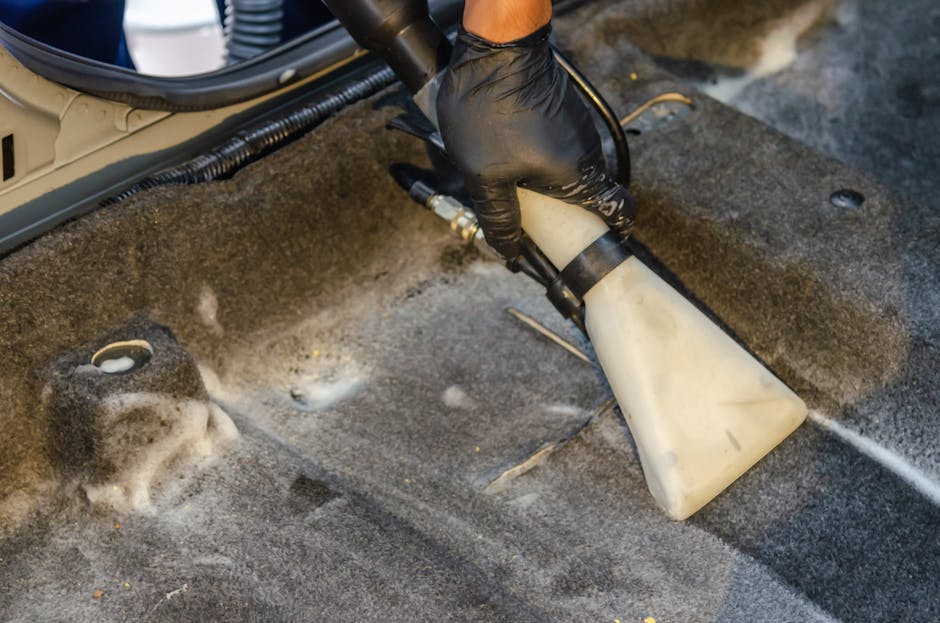 A technician wearing a black glove is using a handheld steam or extraction cleaning device on a stained carpet in a residential or commercial space. The device has a white nozzle and black hose, emitting a stream of cleaning solution or steam onto the dark, textured carpet surface to remove deep-set stains and dirt. Surrounding the area are visible parts of the carpeted floor, with a circular foam or fabric patch, possibly a vent or protective cover, nearby. The scene is well-lit, emphasizing the texture and cleanliness of the carpet area, with some foam residues indicating active deep cleaning. This image illustrates professional surface cleaning and deep cleaning processes performed by Deep Cleaning Highbury to maintain hygienic and well-maintained indoor environments.