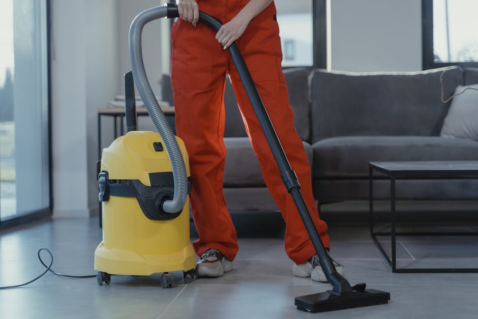 A person dressed in orange overalls is operating a yellow and black vacuum cleaner with a grey hose, cleaning a light grey tiled floor in a modern living room. The room features a dark grey sofa with cushions and a glass-topped coffee table, with natural light coming through large windows in the background. The scene highlights a thorough surface cleaning process aimed at maintaining hygiene and cleanliness within a residential setting, as part of the services offered by Deep Cleaning Highbury.