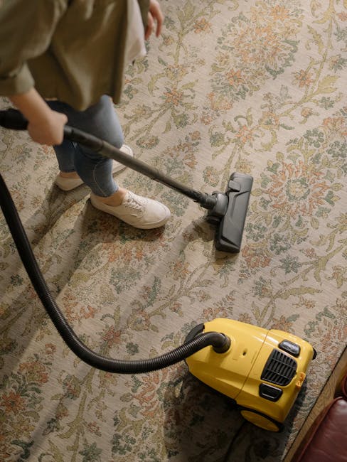 A technician wearing a black glove is using a handheld steam or extraction cleaning device on a stained carpet in a residential or commercial space. The device has a white nozzle and black hose, emitting a stream of cleaning solution or steam onto the dark, textured carpet surface to remove deep-set stains and dirt. Surrounding the area are visible parts of the carpeted floor, with a circular foam or fabric patch, possibly a vent or protective cover, nearby. The scene is well-lit, emphasizing the texture and cleanliness of the carpet area, with some foam residues indicating active deep cleaning. This image illustrates professional surface cleaning and deep cleaning processes performed by Deep Cleaning Highbury to maintain hygienic and well-maintained indoor environments.