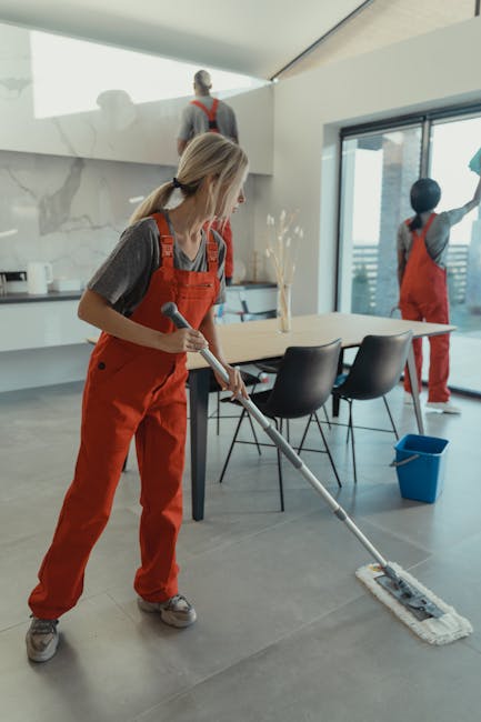 A female cleaner in red overalls and grey top is mopping a large grey tile floor in a modern, well-lit dining area with a large window. In the background, another cleaner in matching red overalls and grey top is cleaning the window sill. The room features a contemporary white marble kitchen island, black chairs, and a blue bucket on the floor. The scene depicts thorough surface cleaning and sanitisation, highlighting attention to hygiene and maintaining a spotless environment. Deep Cleaning Highbury offers domestic cleaning services that focus on detailed cleaning of surfaces, including tiles, floors, and glass, ensuring a pristine and hygienic space.