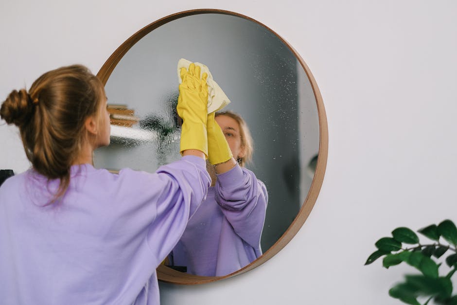 A woman wearing a light purple long-sleeve shirt and yellow rubber cleaning gloves is using a white cloth to wipe a foggy, reflective round mirror mounted on a white wall. The mirror's surface shows water droplets and condensation, indicating a recent cleaning or steam from a bathroom or kitchen area. The background includes a small portion of a wooden shelf with a few stacked books, and a green leafy plant is partially visible in the lower right corner. The image emphasizes surface cleaning and sanitisation, showcasing deep cleaning practices, with natural lighting highlighting the cleanliness and shine of the mirror. Deep Cleaning Highbury’s professional cleaning services focus on maintaining hygiene and spotless results in domestic settings. The overall scene illustrates meticulous surface cleaning and the importance of regular sanitisation in home environments.