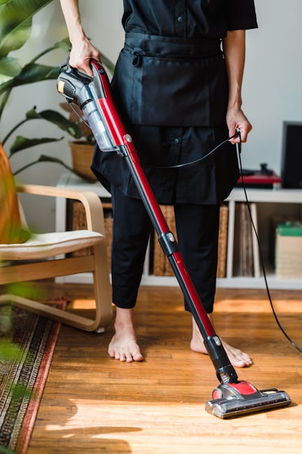 A female cleaner in red overalls and grey top is mopping a large grey tile floor in a modern, well-lit dining area with a large window. In the background, another cleaner in matching red overalls and grey top is cleaning the window sill. The room features a contemporary white marble kitchen island, black chairs, and a blue bucket on the floor. The scene depicts thorough surface cleaning and sanitisation, highlighting attention to hygiene and maintaining a spotless environment. Deep Cleaning Highbury offers domestic cleaning services that focus on detailed cleaning of surfaces, including tiles, floors, and glass, ensuring a pristine and hygienic space.