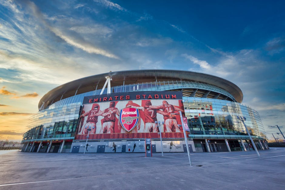 Exterior view of Emirates Stadium, home of Arsenal Football Club, during late afternoon with a partly cloudy sky. The stadium's modern glass facade reflects the surrounding area, and a large digital screen displays Arsenal players in red kits with the club's emblem. The area in front of the stadium is clean, featuring paved walkways with minimal foot traffic and a few street lamps. The lighting emphasizes the sleek curves of the stadium architecture and the vibrant colors on the digital display, exemplifying professional cleaning and maintenance of the venue's exterior surfaces. This image aligns with services offered by Deep Cleaning Highbury for end of tenancy cleaning near Emirates Stadium in Highbury, highlighting the importance of surface cleaning, sanitisation, and exterior maintenance in a commercial sports venue.