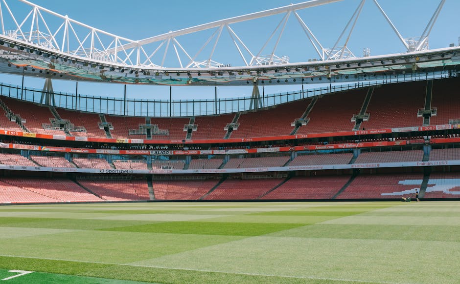 Interior view of a large sports stadium with a well-maintained green grass pitch, empty red seating sections, and a white modern roof structure overhead. The lighting is natural, highlighting the cleanliness and orderly arrangement of the seating and field, suitable for surface cleaning and maintenance purposes. The image reflects a pristine condition, emphasizing hygiene and upkeep, aligned with professional cleaning and sanitisation standards. Deep Cleaning Highbury offers comprehensive cleaning services near Emirates Stadium, Highbury, ensuring the maintenance and sanitation of commercial spaces such as stadiums or large venues.