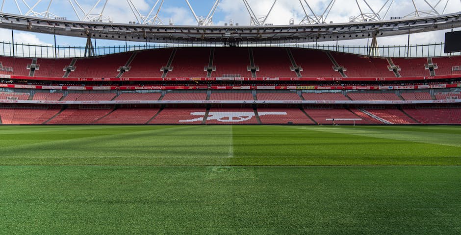 Exterior view of Emirates Stadium, home of Arsenal Football Club, during late afternoon with a partly cloudy sky. The stadium's modern glass facade reflects the surrounding area, and a large digital screen displays Arsenal players in red kits with the club's emblem. The area in front of the stadium is clean, featuring paved walkways with minimal foot traffic and a few street lamps. The lighting emphasizes the sleek curves of the stadium architecture and the vibrant colors on the digital display, exemplifying professional cleaning and maintenance of the venue's exterior surfaces. This image aligns with services offered by Deep Cleaning Highbury for end of tenancy cleaning near Emirates Stadium in Highbury, highlighting the importance of surface cleaning, sanitisation, and exterior maintenance in a commercial sports venue.