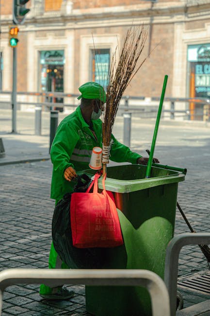 A street cleaner dressed in bright orange high-visibility clothing, including a hood and gloves, is sweeping the pavement with a red broom. Next to them, there is a green trash bin attached to a mobil