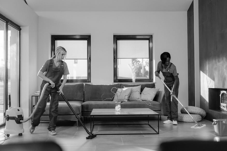 A close-up view of a professional vacuum cleaner with a metallic body and black and orange accents, resting on a wooden floor in a residential room. The vacuum hose is attached, extending towards the 