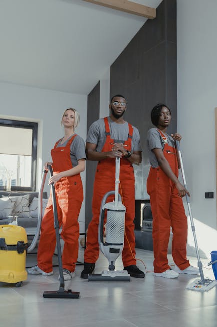 Three professional cleaners from Deep Cleaning Highbury dressed in grey shirts and red overalls are standing inside a modern living room with natural lighting. They are equipped with vacuum cleaners and other cleaning tools, ready for surface cleaning or deep cleaning tasks. The room features a large window, a beige sofa, and a tiled fireplace area, with a polished, dust-free floor surface. The image showcases a professional approach to domestic cleaning, emphasizing hygiene and thoroughness in maintaining a clean and hygienic environment for residential spaces. The overall scene highlights the company's expertise in professional cleaning services in Highbury, suitable for same-day emergency cleaning needs in N5.