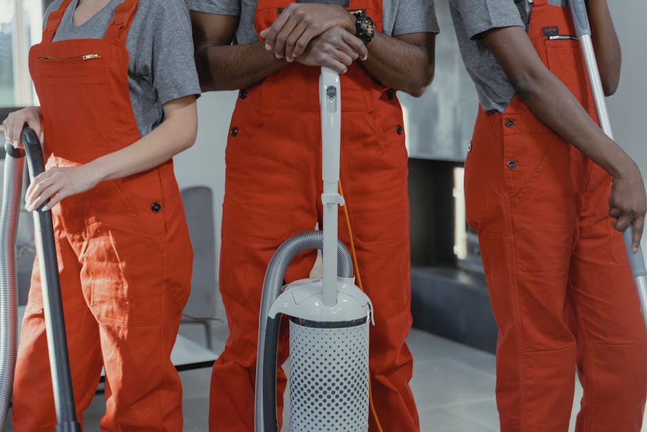 Close-up of three professional cleaners wearing orange overalls, using vacuum cleaners on residential surfaces. The middle cleaner holds a white vacuum with a cylindrical body and a flexible hose, performing surface cleaning in a modern interior. The other two cleaners, partially visible, are also holding vacuum attachments and appear to be engaged in deep cleaning tasks. The environment features smooth, tiled flooring and neutral-colored walls, with bright lighting highlighting the cleanliness of the space. The image emphasizes thorough sanitisation and surface cleaning services provided by Deep Cleaning Highbury, aimed at maintaining hygiene in homes or commercial settings.